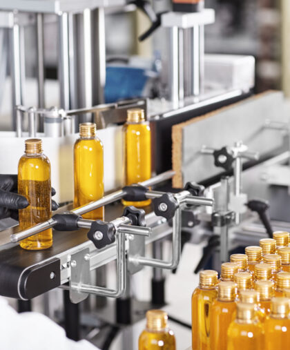 Rear view of unrecognizable factory worker in gown and gloves controlling production process of manufacturing cosmetics at modern laboratory, taking bottle of golden yellow liquid from conveyor line Rear view of unrecognizable factory worker in gown and gloves controlling production process of manufacturing cosmetics at modern laboratory, taking bottle of golden yellow liquid from conveyor line